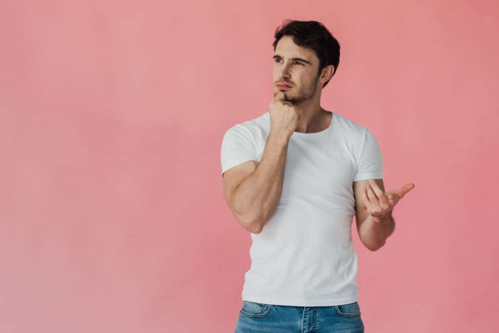 pensive muscular man in white t-shirt propping face with fist and counting on fingers isolated on pink